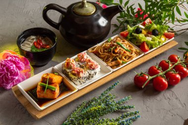 Tam öğle yemeği. Yunan salatası, cam erişte, rulo, somon filetosu, erişte çorbası ve gri zemin üzerinde çay olan çaydanlık..