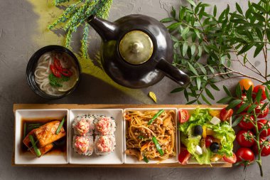 Tam öğle yemeği. Yunan salatası, cam erişte, rulo, somon filetosu, erişte çorbası ve gri zemin üzerinde çay olan çaydanlık..