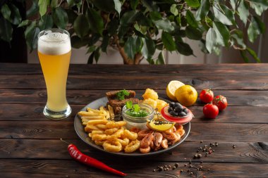 Bir bardak köpüklü bira ve abur cuburlu bir tabak: karides, kalamar halkaları, patates kızartması, çavdar ekmeği, ahşap zemin üzerinde zeytin.