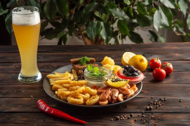 Bir bardak köpüklü bira ve abur cuburlu bir tabak: karides, kalamar halkaları, patates kızartması, çavdar ekmeği, ahşap zemin üzerinde zeytin.