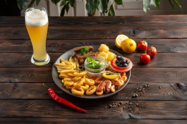 Bir bardak köpüklü bira ve abur cuburlu bir tabak: karides, kalamar halkaları, patates kızartması, çavdar ekmeği, ahşap zemin üzerinde zeytin.