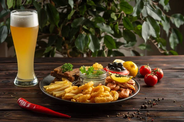 Bir bardak köpüklü bira ve abur cuburlu bir tabak: karides, kalamar halkaları, patates kızartması, çavdar ekmeği, ahşap zemin üzerinde zeytin.