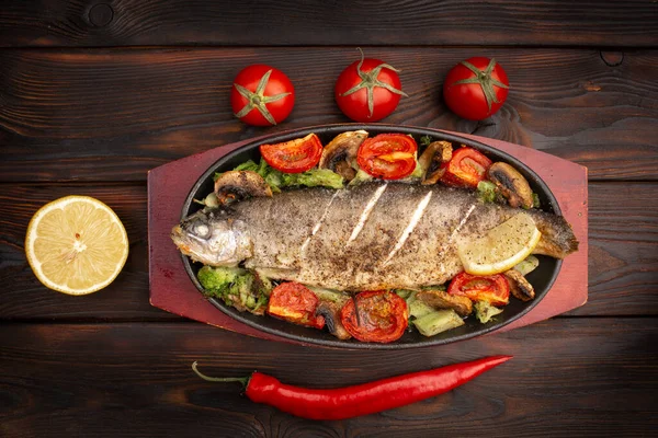 baked trout with vegetables on an oval plate decorated with fresh tomatoes and chili peppers on a dark wooden background