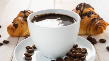 Beyaz ahşap bir masada iki kruvasan ve bir fincan kahve.