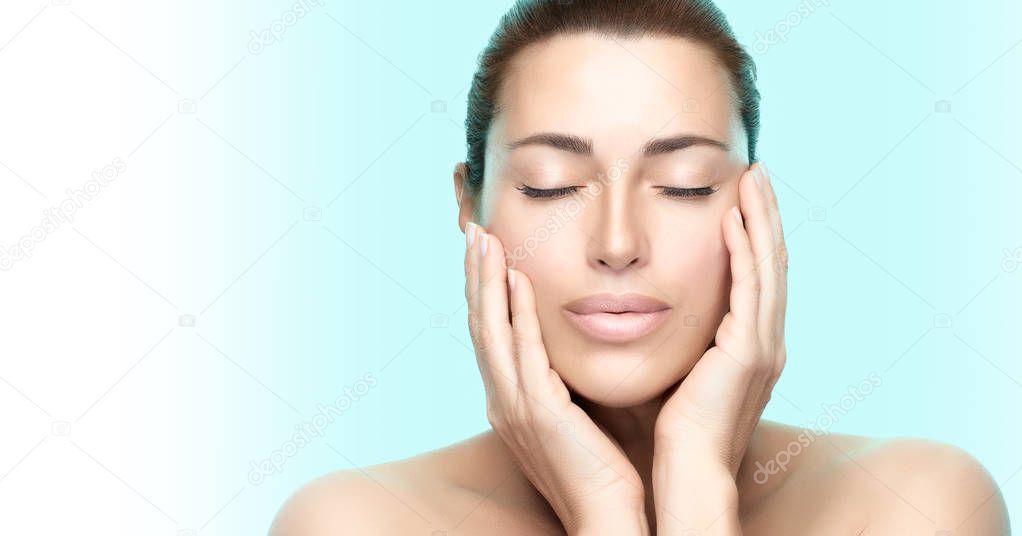 Beauty woman face. Beautiful brunette model girl with clean fresh skin and serene expression holding her hands to cheeks with closed eyes on blue and white background. Spa, Beauty and Skincare concept