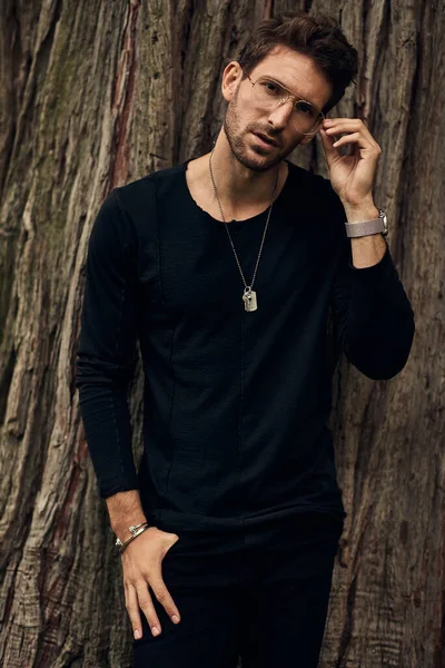 Handsome man in black standing agains tree and touching his glas — Stockfoto