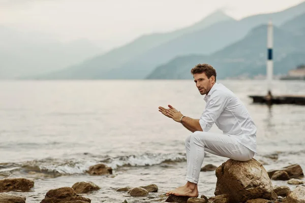 Modelo masculino guapo se sienta en un fondo de lago y montañas — Foto de Stock
