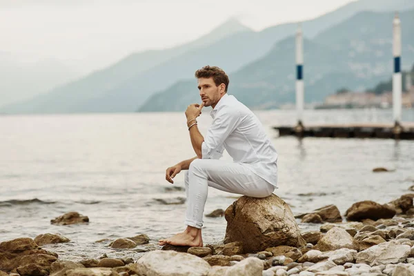 Modelo masculino guapo se sienta en un fondo de lago y montañas — Foto de Stock