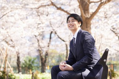 Japon bir erkek işadamı yüzünde bir gülümsemeyle bir bankta oturuyor.