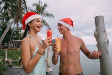 Genç bir çift, tropik bir adada Noel şapkası takıp okyanusta kokteyl içiyor.. 