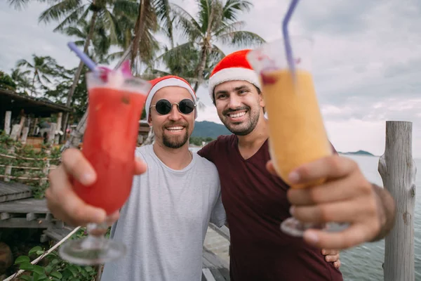 Молодая пара геев с коктейлями в рождественских шапках на океане — стоковое фото