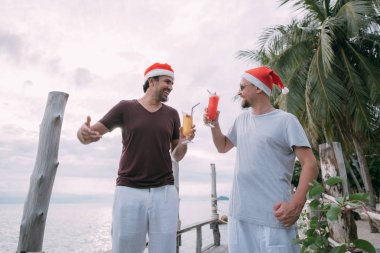 Noel şapkalarında kokteylleri olan arkadaşlar okyanusta tropi üzerinde