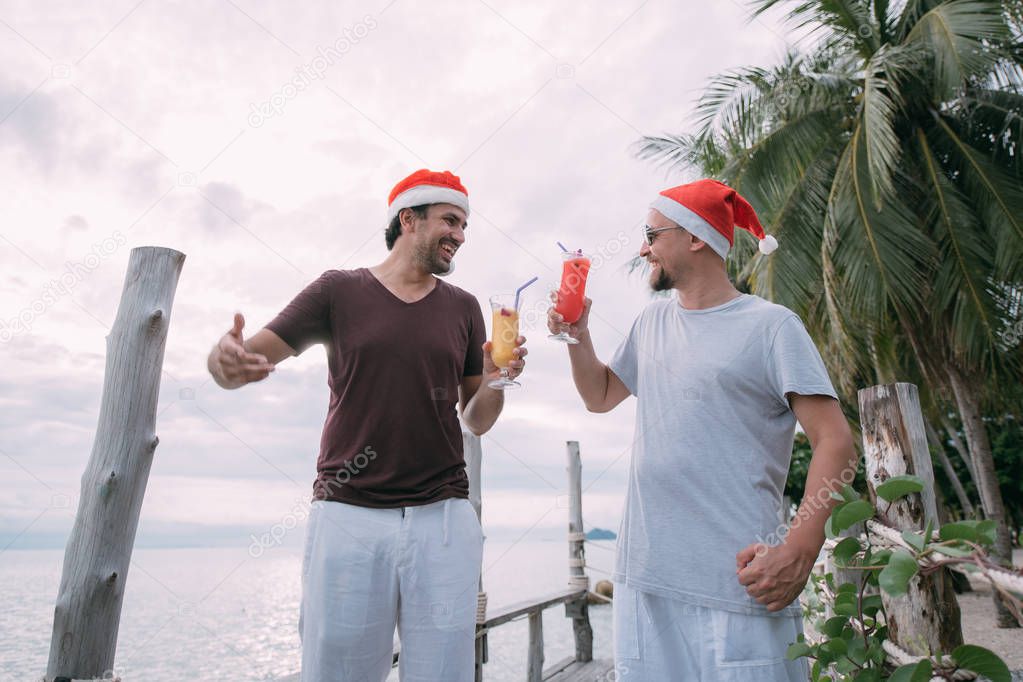 Friends with cocktails in Christmas caps on the ocean on a tropi