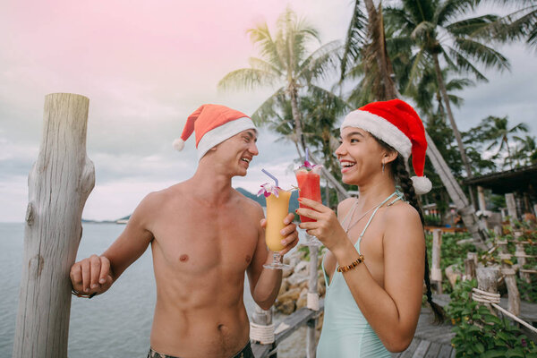 Young couple with cocktails in Christmas caps on the ocean on a tropical island. 