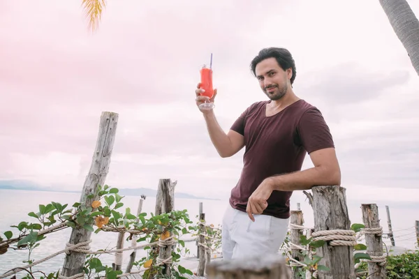 A young man stands by the sea on a wooden bridge with a cocktail