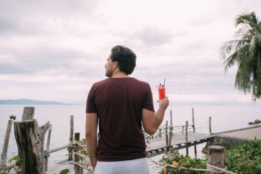Genç bir adam, elinde bir kokteyl ile, tahta bir köprüde, denizin kenarında duruyor.