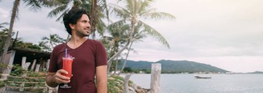 Genç bir adam, elinde bir kokteyl ile, tahta bir köprüde, denizin kenarında duruyor.