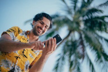 Memnun bir adam elinde telefonla palmiye ağaçlarının arka planında dikiliyor.. 