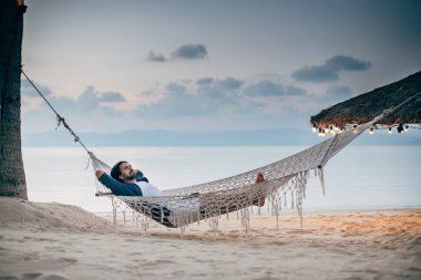 Bir adam sükunetten hoşlanır, okyanusun arka planında bir hamakta yatar ve gün batımını seyreder.. 