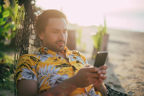 Een jonge man ligt in een hangmat op een zonnig strand met een telefoon. — Stockfoto