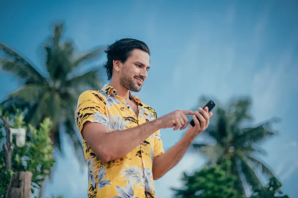 A contented man is standing with a phone in his hands on a background of palm trees.