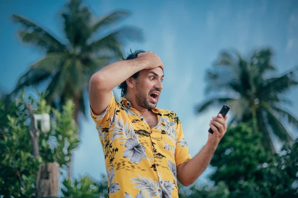 An upset man stands with a phone in his hands on a background of palm trees.