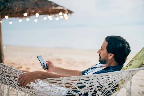 Een man ligt in een hangmat met een telefoon op de achtergrond van de oceaan en zonsondergang. — Stockfoto