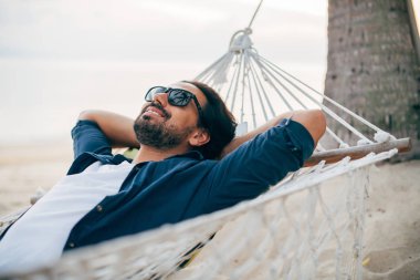 Bir adam sükunetten hoşlanır, okyanusun arka planında bir hamakta yatar ve gün batımını seyreder.