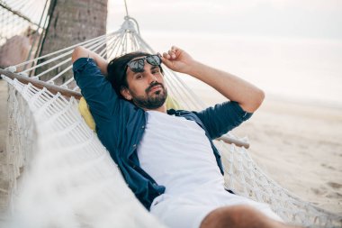 Bir adam sükunetten hoşlanır, okyanusun arka planında bir hamakta yatar ve gün batımını seyreder.