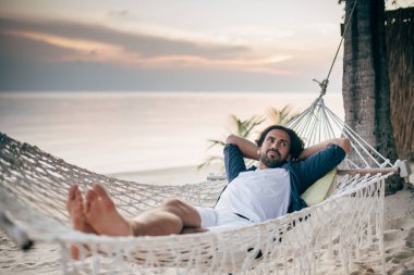 Bir adam sükunetten hoşlanır, okyanusun arka planında bir hamakta yatar ve gün batımını seyreder.. 