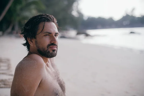 A handsome man lies on the sand by the ocean.