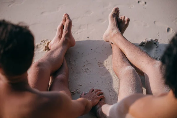 Dos hombres gay están sentados en la arena junto al océano, tomados de la mano . — Foto de Stock