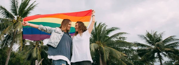 Dwie kobiety z tęczową flagą na plaży na tle palm — Zdjęcie stockowe