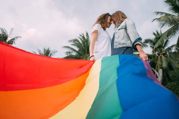 Dwie kobiety z tęczową flagą na plaży na tle palm — Zdjęcie stockowe