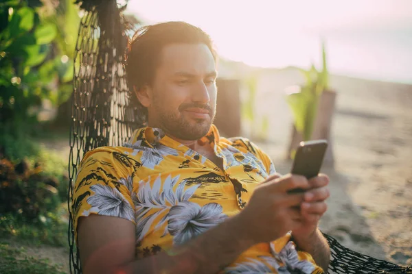 Een jonge man ligt in een hangmat op een zonnig strand met een telefoon. — Stockfoto