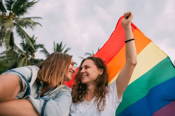 Dwie kobiety z tęczową flagą na plaży na tle palm — Zdjęcie stockowe