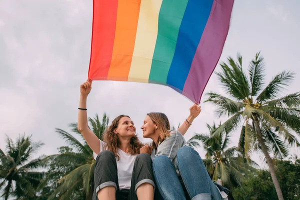 Dwie kobiety z tęczową flagą na plaży na tle palm — Zdjęcie stockowe