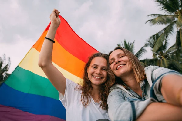Two women with rainbow flag on the beach on a background of palm trees — ストック写真