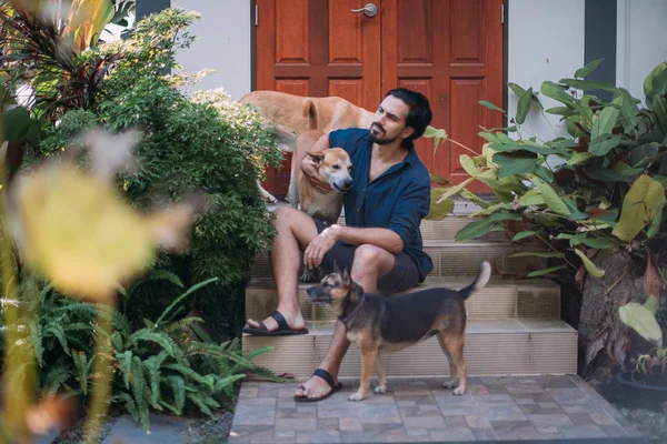 Ein Mann mit Hunden sitzt auf der Veranda eines Hauses in einem tropischen Garten — Stockfoto