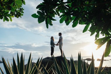 Bir adam ve bir kız gün batımında deniz kıyısında büyük bir taşın üzerinde dururlar.