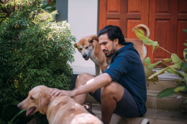Köpekli bir adam tropik bir bahçedeki bir evin verandasında oturur.