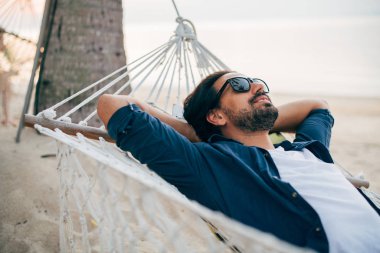 Bir adam sükunetten hoşlanır, okyanusun arka planında bir hamakta yatar ve gün batımını seyreder..