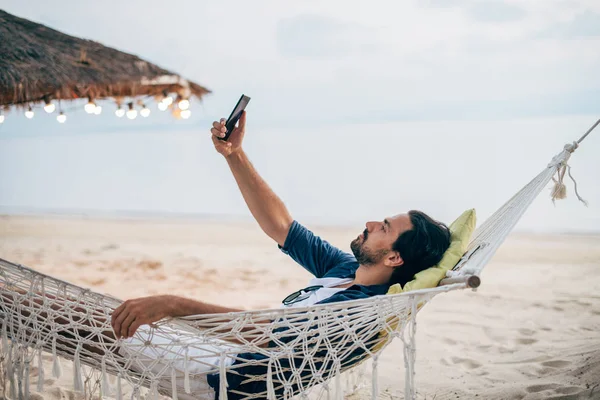 A man  lies in a hammock with a phone on the background of the ocean and sunset.