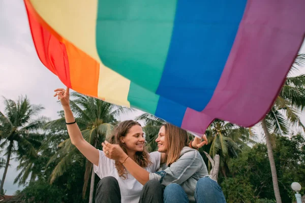 Twee vrouwen met regenboogvlag op het strand op een palmachtergrond — Stockfoto