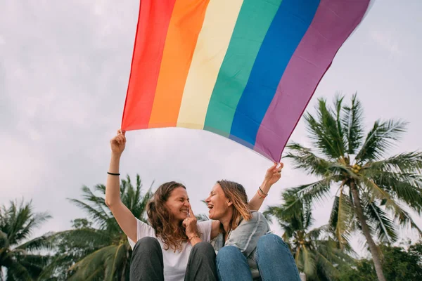 Dos mujeres con bandera de arco iris en la playa sobre un fondo de palma — Foto de Stock