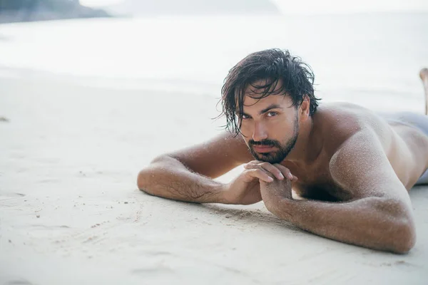 A handsome man lies on the sand by the ocean.