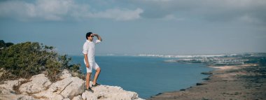 Denize bakan kayalıklarda bir adam. Beyaz elbiseli yakışıklı adam kayalık sahildeki okyanus manzarasının tadını çıkarıyor. # Denizin ötesindeki genç adam #