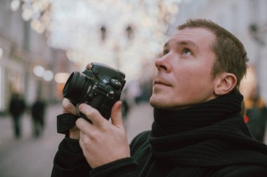 Elinde kamera olan bir adam kışın eski şehrin caddesini fotoğraflıyor. Arka planda sanatsal dekore edilmiş sokak lambalarının altında kamera ve eşarplı bir fotoğrafçı.