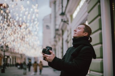 Elinde kamera olan bir adam kışın eski şehrin caddesini fotoğraflıyor. Arka planda sanatsal dekore edilmiş sokak lambalarının altında kamera ve eşarplı bir fotoğrafçı.