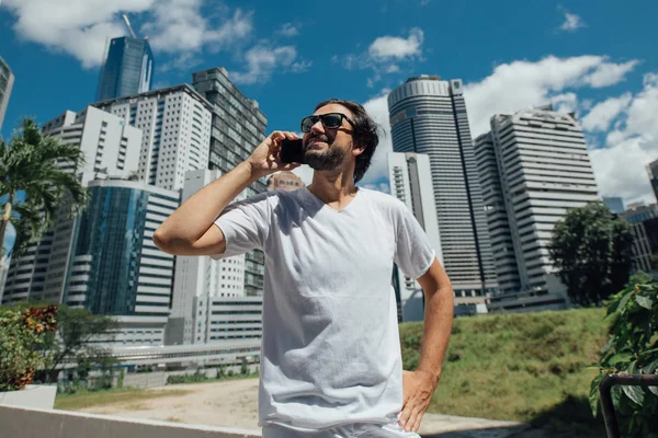 A man talks on the phone against the backdrop of skyscrapers. A young guy in a metropolis writes a message. A tourist walks and talks online in the big city during the day.